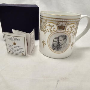 May include: A white ceramic mug with gold detailing, featuring a portrait of a couple. The mug has a handle and is decorated with gold patterns and a crown. Includes a navy box and a Royal Worcester tag.