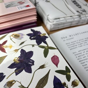 May include: A collection of stationery featuring pressed purple and red columbine flowers on white paper, accompanied by a printed letter titled The Wildflower, and stacks of pink and white envelopes with floral postage stamps.