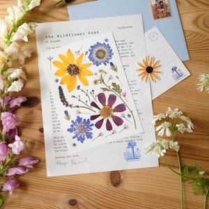 May include: A white card featuring pressed yellow, purple, and blue wildflowers sits atop a letter titled The Wildflower Post dated 03/26/2026. A light blue envelope with a postage stamp, a small flower sticker, and a fountain pen are also present.