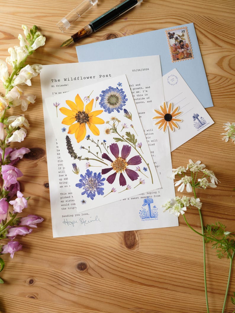 May include: A white card featuring pressed yellow, purple, and blue wildflowers sits atop a letter titled The Wildflower Post dated 03/26/2026. A light blue envelope with a postage stamp, a small flower sticker, and a fountain pen are also present.