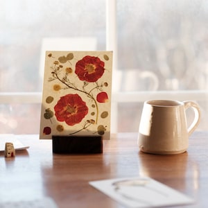 May include: A rectangular card featuring pressed red flowers and green foliage on a cream background, held upright by a dark brown wooden block stand. Beside the stand sits a plain, light-coloured ceramic mug with a handle on a wooden surface.