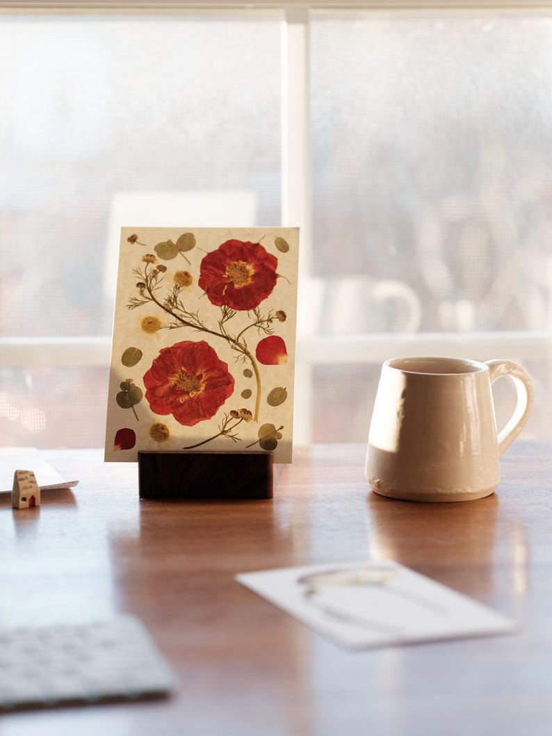 May include: A rectangular card featuring pressed red flowers and green foliage on a cream background, held upright by a dark brown wooden block stand. Beside the stand sits a plain, light-coloured ceramic mug with a handle on a wooden surface.
