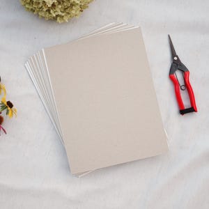 May include: A stack of light tan cardstock paper with white edges. A pair of red and black handled scissors rests on the right side of the image. Dried flowers are visible in the background.
