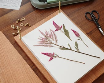 May include: A wooden flower press featuring two light brown rectangular boards held together by four brass bolts and wing nuts. A stack of white paper sits between the boards, displaying three pressed pink celosia flowers.