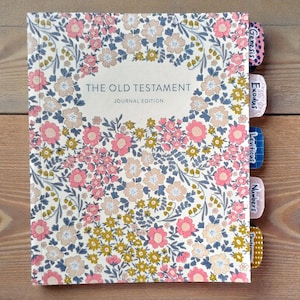 May include: A floral-patterned journal titled "The Old Testament Journal Edition." The cover features a cream background with pink, blue, and yellow flowers. Tabs on the side are labeled with book names like Genesis and Exodus.