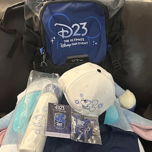 May include: A blue and gray backpack with the text "D23 The Ultimate Disney Fan Event" and the logo for D23. The backpack is surrounded by other Disney merchandise, including a white baseball cap with a Mickey Mouse design, a blue t-shirt with a Mickey Mouse design, and a D23 pin.