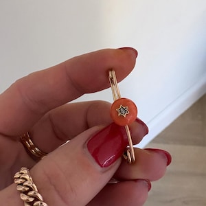 Puede incluir: Broche dorado con una pieza central circular de color coral. El centro tiene un diseño en forma de estrella con una pequeña piedra transparente. El broche lo sostiene una persona con esmalte de uñas rojo y un anillo de cadena dorada.