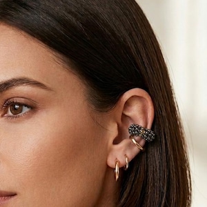 May include: A woman with short, dark brown hair wearing a black ear cuff and three gold hoop earrings. The ear cuff is adorned with small, dark embellishments. The earrings are simple and elegant.