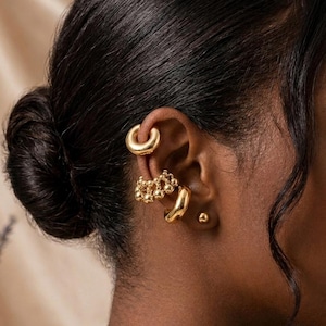 May include: A collection of gold-toned earrings adorns an ear. The earrings include a curved ear cuff, a cluster of small beaded earrings, a small hoop, and a stud. The person's hair is styled in a bun.