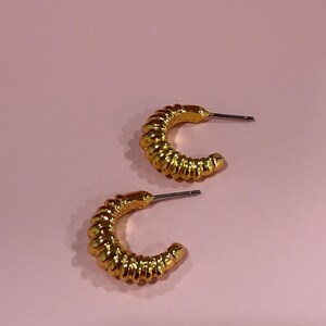 May include: A pair of gold-colored hoop earrings with a textured, ridged design. The earrings are crescent-shaped and have a post for insertion. The background is a solid, light pink color.