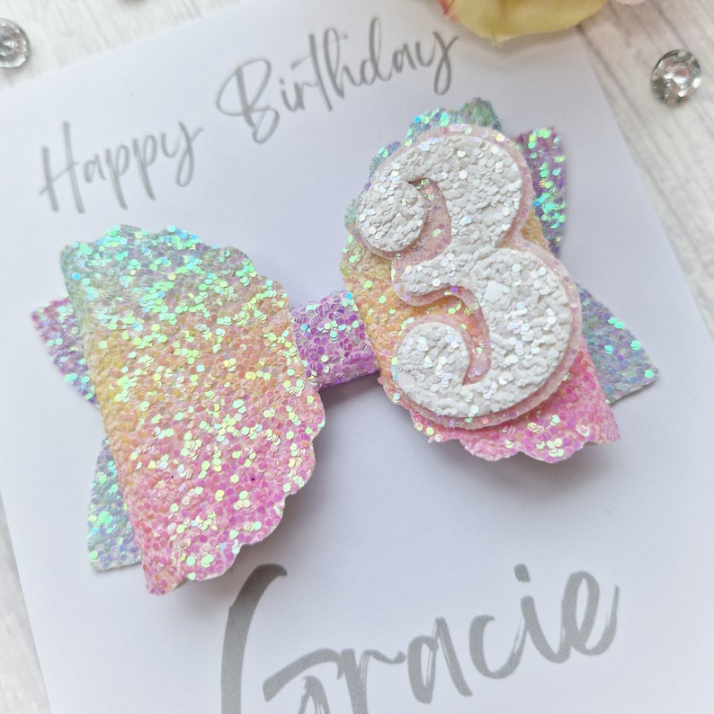 May include: A rainbow glitter bow with a white glitter number 3 on top. The bow is on a white card with the text "Happy Birthday Gracie".