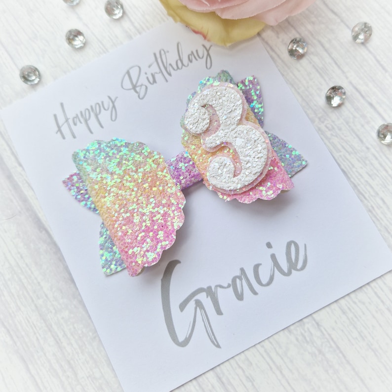 May include: A white card with the text "Happy Birthday" and "Gracie" written in gray. A sparkly iridescent pink and blue bow with a white number 3 is attached to the card.