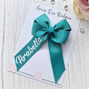 May include: A teal green hair bow with a white personalized name tag that says "Arabella". The bow is on a white card with the text "Gracie Rose Boutique" and social media handles.