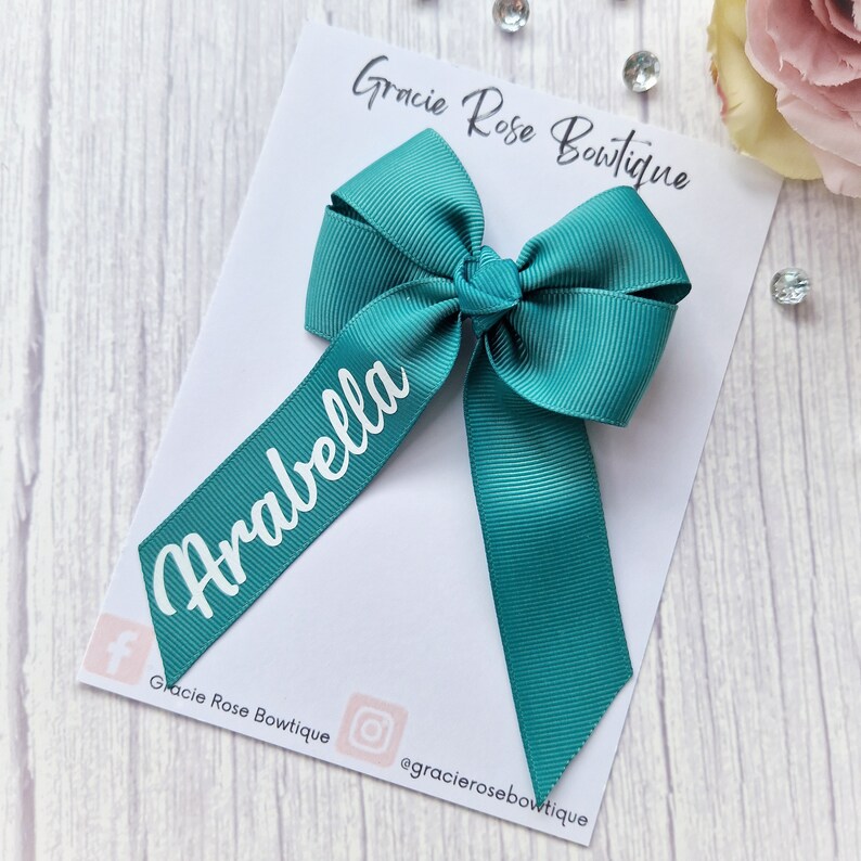 May include: A teal green hair bow with a white personalized name tag that says "Arabella". The bow is on a white card with the text "Gracie Rose Boutique" and social media handles.