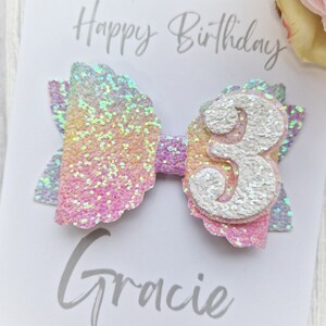 May include: A pink and iridescent glitter bow with a white glitter number 3 on top. The bow is on a white card with the text "Happy Birthday Gracie" written on it.