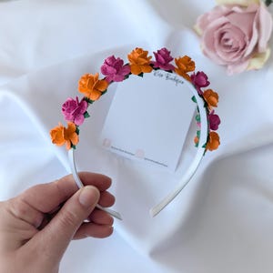 May include: A white headband decorated with bright orange and fuchsia paper flowers. The floral crown features green leaves. A hand holds the headband, and a blank white card is visible. The background is a white, silky fabric.