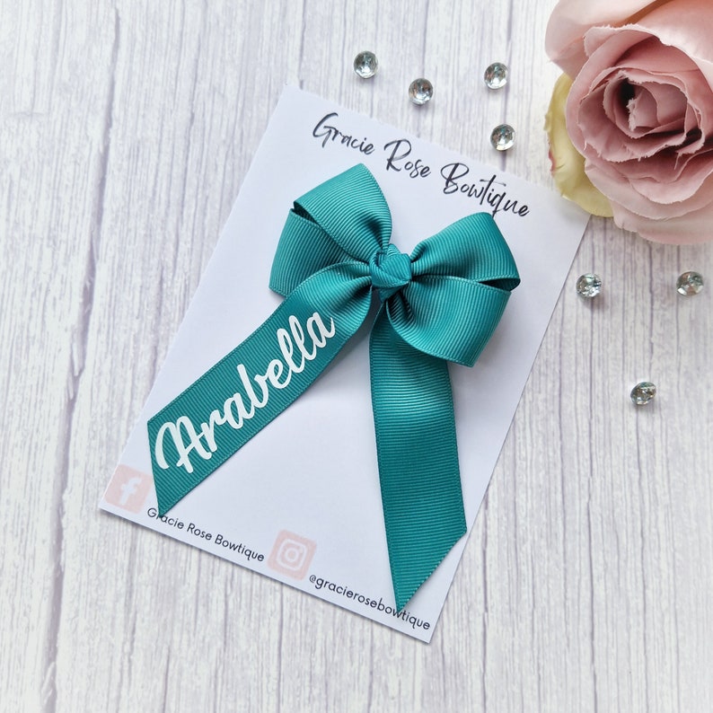 May include: Teal personalized hair bow with the name "Arabella" in white lettering. The bow is on a white card with the text "Gracie Rose Boutique" and social media handles.