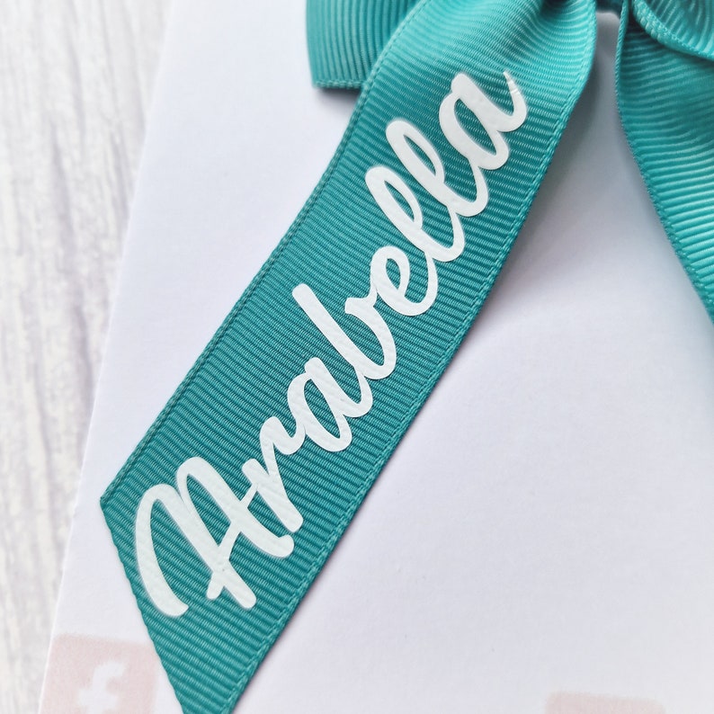 May include: A teal ribbon with the name "Arabella" printed in white cursive lettering.