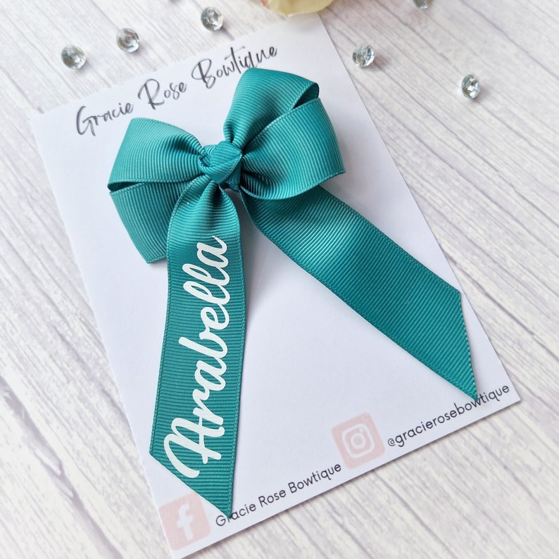 May include: Teal hair bow with the name "Arabella" printed in white on the ribbon. The bow is on a white card with the text "Gracie Rose Boutique" and social media handles.