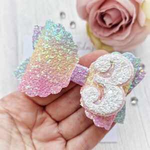 May include: A rainbow glitter bow with a white glitter number 3 attached. The bow is made of iridescent glitter fabric.