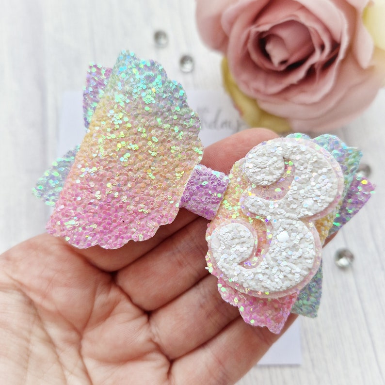 May include: A rainbow glitter bow with a white glitter number 3 attached. The bow is made of iridescent glitter fabric.