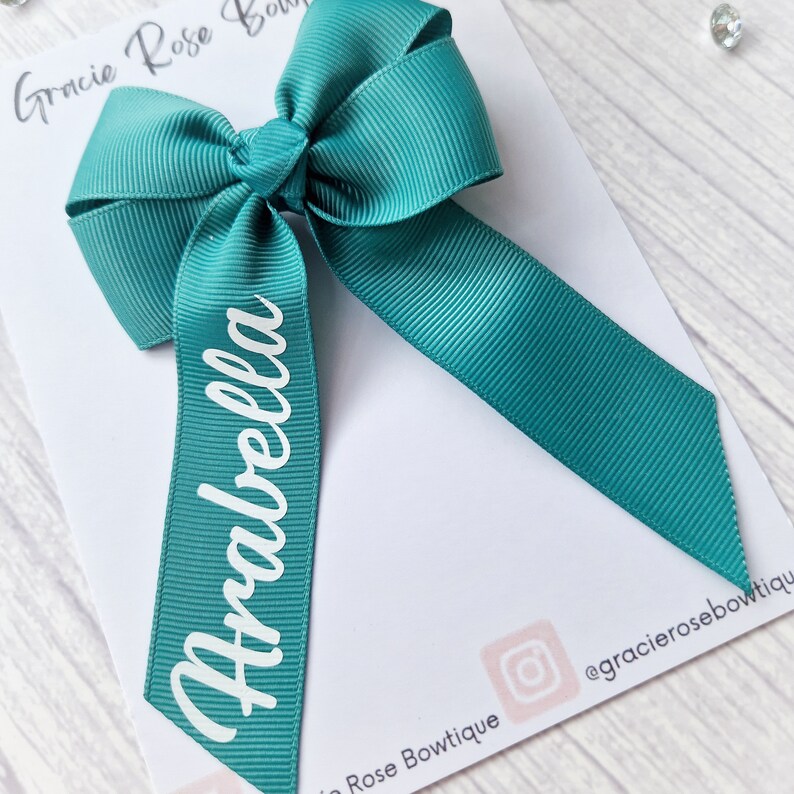 May include: Teal blue ribbon bow with the name Arabella written in white on the tail. The bow is on a white background.