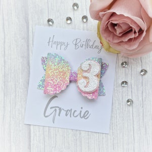 May include: A rainbow glitter hair bow with a white number 3 and the name Gracie on a white card with the text "Happy Birthday".