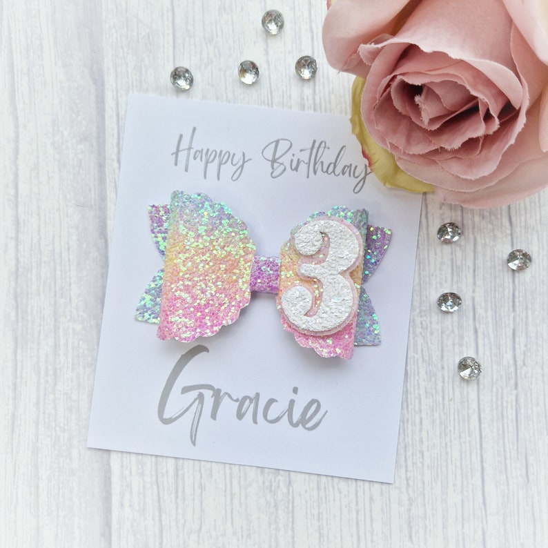 May include: A rainbow glitter hair bow with a white number 3 and the name Gracie on a white card with the text "Happy Birthday".