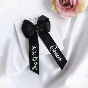 May include: Black ribbon hair bow with the text "Class Of 2026" and "Gracie" in white script. The bow is on a white card, with a pink rose in the background. The bow is made of a grosgrain ribbon.