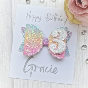 May include: A white card with the text "Happy Birthday Gracie" and a glittery rainbow bow with a white number 3 on top.