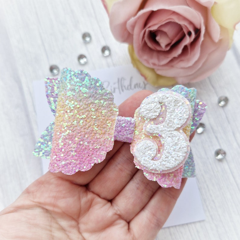 May include: A rainbow glitter bow with a white glitter number 3. The bow is made of iridescent glitter fabric and has a scalloped edge.