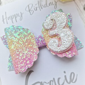May include: A rainbow glitter bow with a white glitter number 3 in the center. The bow is on a white card with the text "Happy Birthday" written in black.