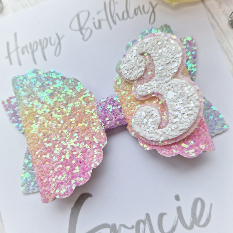 May include: A rainbow glitter bow with a white glitter number 3 in the center. The bow is on a white card with the text "Happy Birthday" written in black.