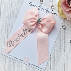 May include: A pink hair bow with a silver glitter name "Arabella" on a white card with the text "Gracie Rose Boutique" and the social media handle "@gracieroseboutique".