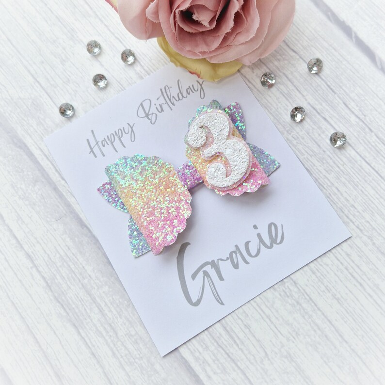 May include: A white card with the text "Happy Birthday" and the name "Gracie" written in gray. A rainbow glitter bow with a white number 3 is attached to the card.