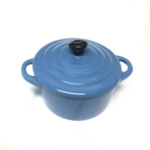 May include: A light blue enameled cast iron Dutch oven with a matching lid and two side handles. The lid features a circular ridged design and a small, smooth, black knob handle centered on top. The pot has a glossy finish and rounded sides.