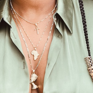 May include: Three gold necklaces with different pendant designs: an Ankh cross, the African continent, and a bust of Nefertiti. The necklaces are layered and worn over a light green collared shirt. A black braid with decorative beads is visible on the right.