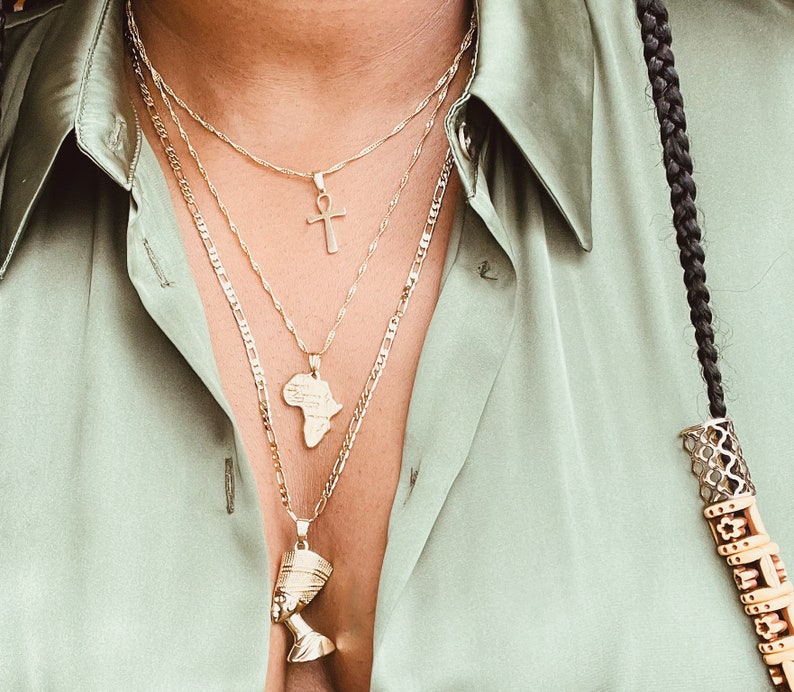May include: Three gold necklaces with different pendant designs: an Ankh cross, the African continent, and a bust of Nefertiti. The necklaces are layered and worn over a light green collared shirt. A black braid with decorative beads is visible on the right.