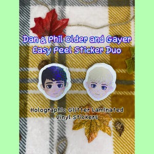 May include: Two holographic glitter laminated vinyl stickers featuring cartoon illustrations of Dan and Phil. The text "Dan & Phil Older and Gayer Easy Peel Sticker Duo" is printed above the stickers.
