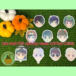 May include: A selection of holographic reflect vinyl stickers with chibi-style character illustrations in costumes. The stickers are displayed on a green surface with pumpkins. Text reads "Dan and Phil Spooky Week Edition Chibis".