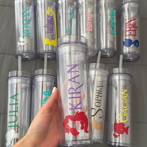May include: Clear, personalized water bottles with silver lids and straws. Each bottle features a name in a different color and a matching character graphic. The names include RUMAN, Julieta, Kiran, Sophia, and others. The bottles are cylindrical and appear to be made of plastic.