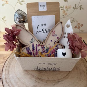 May include: A gift basket on a wooden slice, containing a wooden butterfly, a tea infuser, a tea bag, a small candle with a heart, and dried flowers. The basket has the text "HIP HIP hurra" and "VON HERZEN".