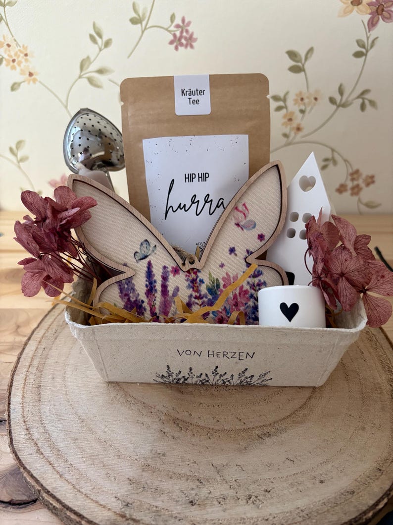 May include: A gift basket on a wooden slice, containing a wooden butterfly, a tea infuser, a tea bag, a small candle with a heart, and dried flowers. The basket has the text "HIP HIP hurra" and "VON HERZEN".