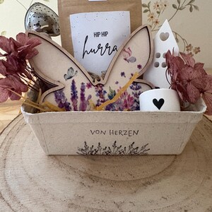 May include: A gift basket with a bunny ear decoration, a tea bag, and a small white candle holder with a heart. The basket has the text "VON HERZEN" and is filled with dried flowers and a tea infuser.