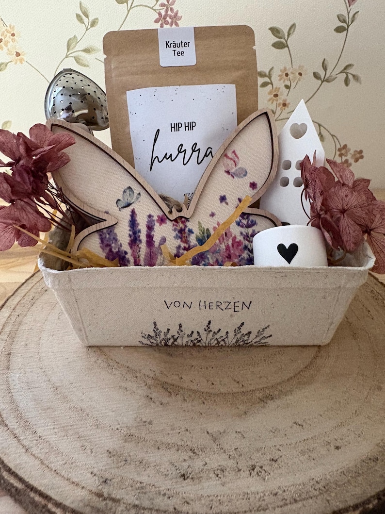 May include: A gift basket with a bunny ear decoration, a tea bag, and a small white candle holder with a heart. The basket has the text "VON HERZEN" and is filled with dried flowers and a tea infuser.
