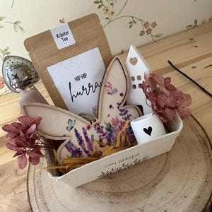May include: A decorative gift basket on a wooden slice, containing a tea bag, a bunny-shaped wooden ornament with floral designs, a small white house, a candle with a heart, and dried flowers. The tea bag has the text "HIP HIP hurra".