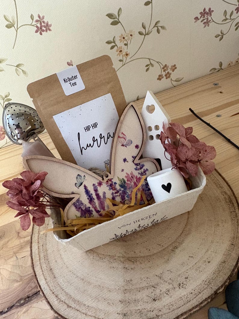 May include: A decorative gift basket on a wooden slice, containing a tea bag, a bunny-shaped wooden ornament with floral designs, a small white house, a candle with a heart, and dried flowers. The tea bag has the text "HIP HIP hurra".