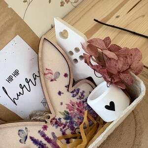 May include: A decorative Easter-themed gift set in a wooden tray. Includes a wooden bunny with floral print, a small white cup with a black heart, a white heart-shaped decoration, dried flowers, and a card with the text "HIP HIP hurra".