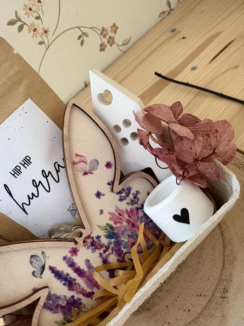 May include: A decorative Easter-themed gift set in a wooden tray. Includes a wooden bunny with floral print, a small white cup with a black heart, a white heart-shaped decoration, dried flowers, and a card with the text "HIP HIP hurra".