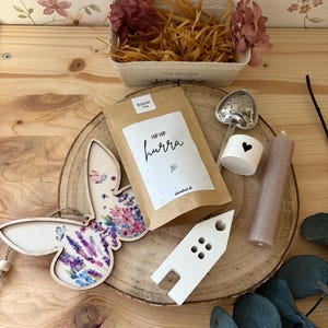 May include: A collection of decorative items on a wooden surface. Includes a butterfly-shaped wooden ornament with a floral design, a bag of tea with the text "HIP HIP hurra", a heart-shaped tea infuser, a candle, and a small white house-shaped object.